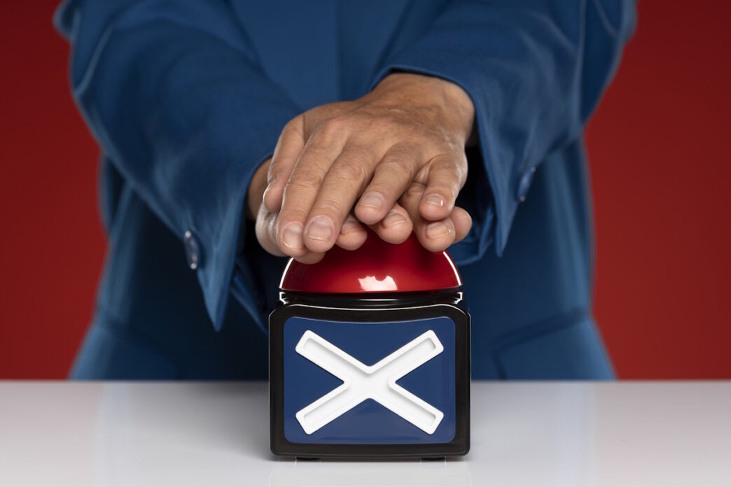 A person wearing a blue suit presses a red buzzer with a large white 'X' on a black box, symbolizing rejection or prohibition